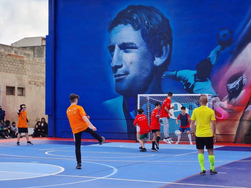 Ragazzi che giocano a calcio nel campetto di Exmè davanti al murale di Gigi Riva